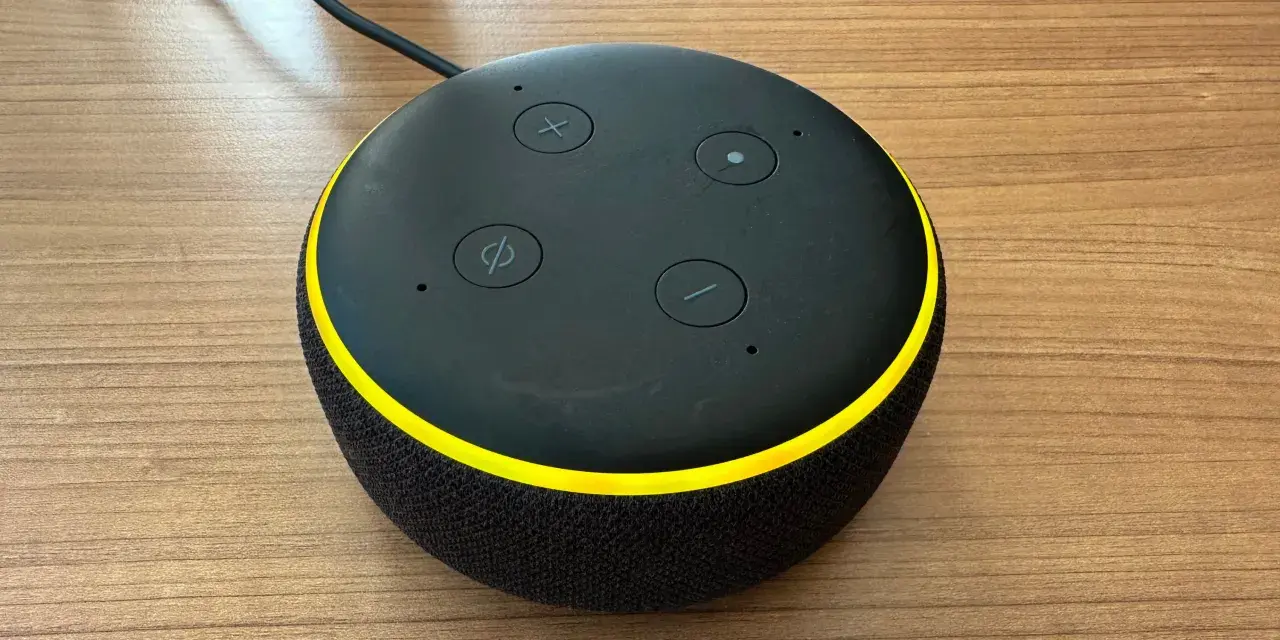 Alexa blinkt gelb. Der smarte Lautsprecher mit schwarzem Stoffbezug und gelbem Ring steht auf einem Holztisch.