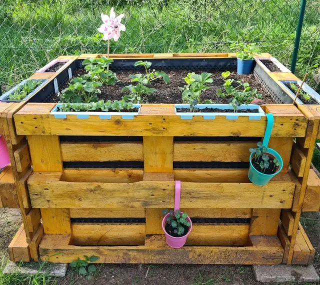 Ein schmales Hochbeet aus Paletten, bepflanzt mit Erdbeeren und Kräutern, geschmückt mit bunten Töpfen und einem bunten Windrad.