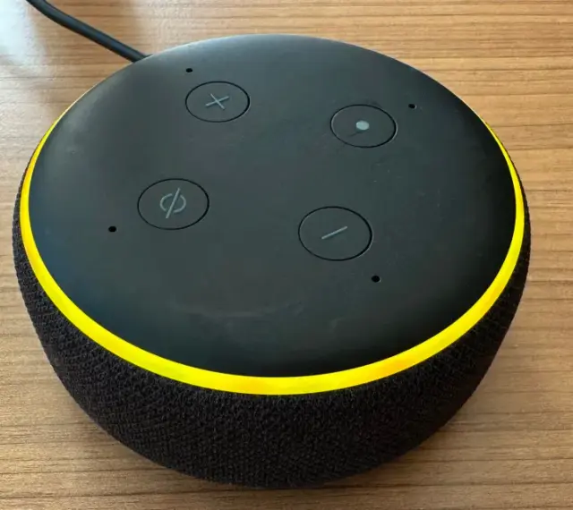 Alexa blinkt gelb. Der smarte Lautsprecher mit schwarzem Stoffbezug und gelbem Ring steht auf einem Holztisch.